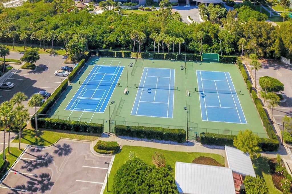 An aerial view of a tennis court complex with multiple courts, surrounded by greenery and a parking area.