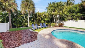 A swimming pool with a curved edge, surrounded by a white fence, palm trees, and a grassy area with Adirondack chairs. - Newly-Renovated, Dog-Friendly Home w Private Heated Pool & Streaming Services (Anna Maria)