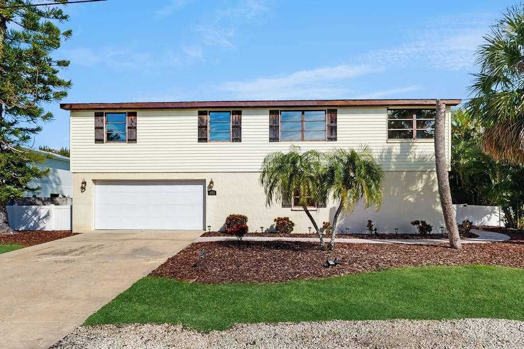Une maison à deux étages avec un garage, un petit jardin devant avec un palmier, et un ciel dégagé.