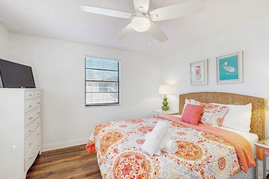 Un dormitorio con cama, ventilador de techo, cómoda, ventana con persianas y dos cuadros en la pared.