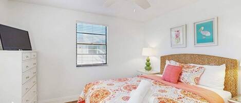 Un dormitorio con cama, ventilador de techo, cómoda, ventana con persianas y dos cuadros en la pared.