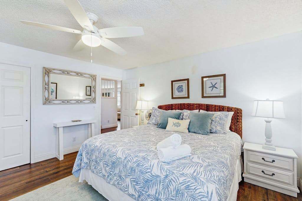 A bedroom with a bed, a ceiling fan, a bedside table, a nightstand, and a mirror.
