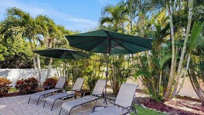 A poolside area with lounge chairs and umbrellas, surrounded by palm trees. - Newly-Renovated, Dog-Friendly Home w Private Heated Pool & Streaming Services (Anna Maria)