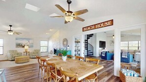 Un'ampia zona giorno con tavolo da pranzo, sedie e una ventola a soffitto. Un cartello recita 'BEACH HOUSE'.