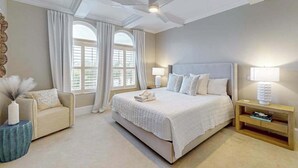 A bedroom with a large bed, a ceiling fan, a nightstand with a lamp, a chair, and a window with curtains. - Oceanview getaway with pool & private patio - steps from the beach (Holmes Beach)