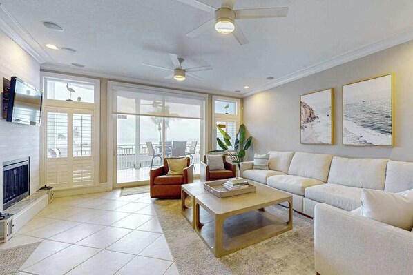 A spacious living room with a fireplace, a ceiling fan, and a large window. - Oceanview getaway with pool & private patio - steps from the beach (Holmes Beach)