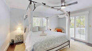 Camera da letto con letto a baldacchino, ventilatore a soffitto e vista sull'esterno attraverso ampie finestre.