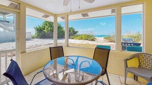 Une pièce lumineuse, baignée de soleil, avec une table en verre, des chaises, un ventilateur de plafond et de grandes fenêtres donnant sur la plage.