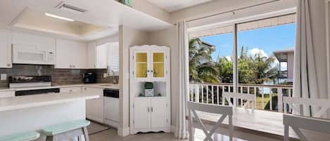 A modern kitchen with white cabinets, a microwave, and a coffee maker. There is a balcony with white chairs and a view of palm trees.