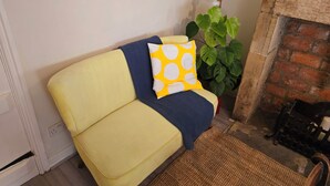 A yellow sofa with a blue blanket and a yellow pillow with white circles, positioned next to a green plant and a brick fireplace. - Cosy Cottage with river stream in Matlock (Matlock)