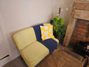 A yellow sofa with a blue blanket and a yellow pillow with white circles, positioned next to a green plant and a brick fireplace. - Cosy Cottage with river stream in Matlock (Matlock)