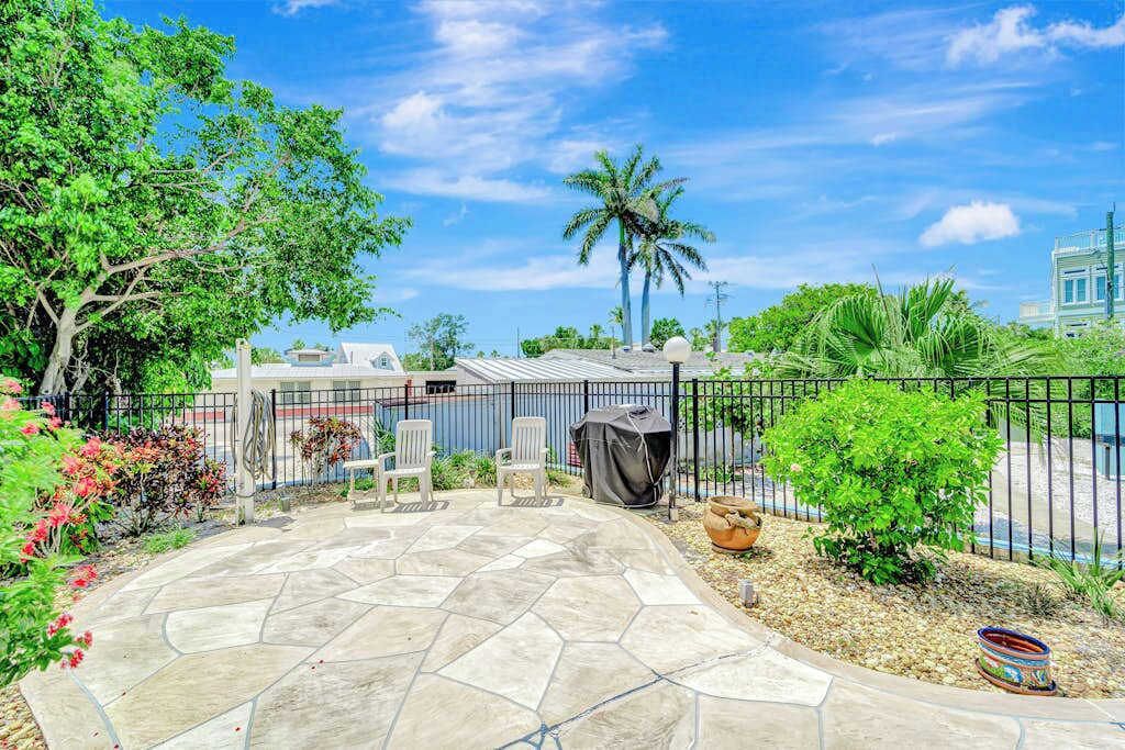 Eine Terrasse mit Steinboden, einem schwarzen Metallzaun, weißen Stühlen und einer Palme.