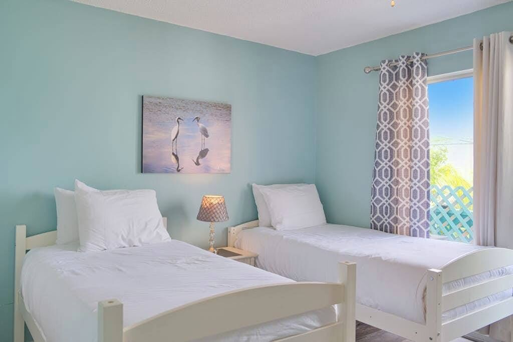 Una habitación con dos camas individuales, una mesita de noche con lámpara, un cuadro de aves colgado en la pared y una ventana con cortina estampada.