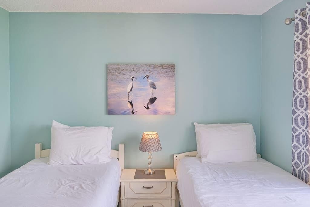 Una habitación con dos camas, una mesita de noche con lámpara y un cuadro de pájaros en la pared.