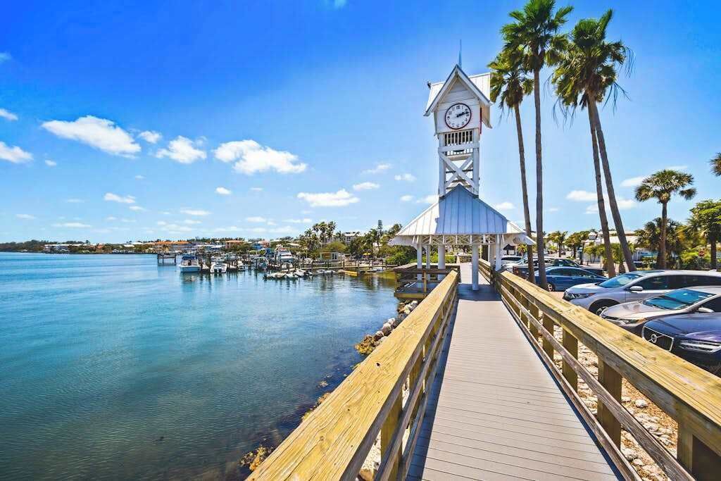 A pier leading to a white building with a clock tower, surrounded by water and palm trees.