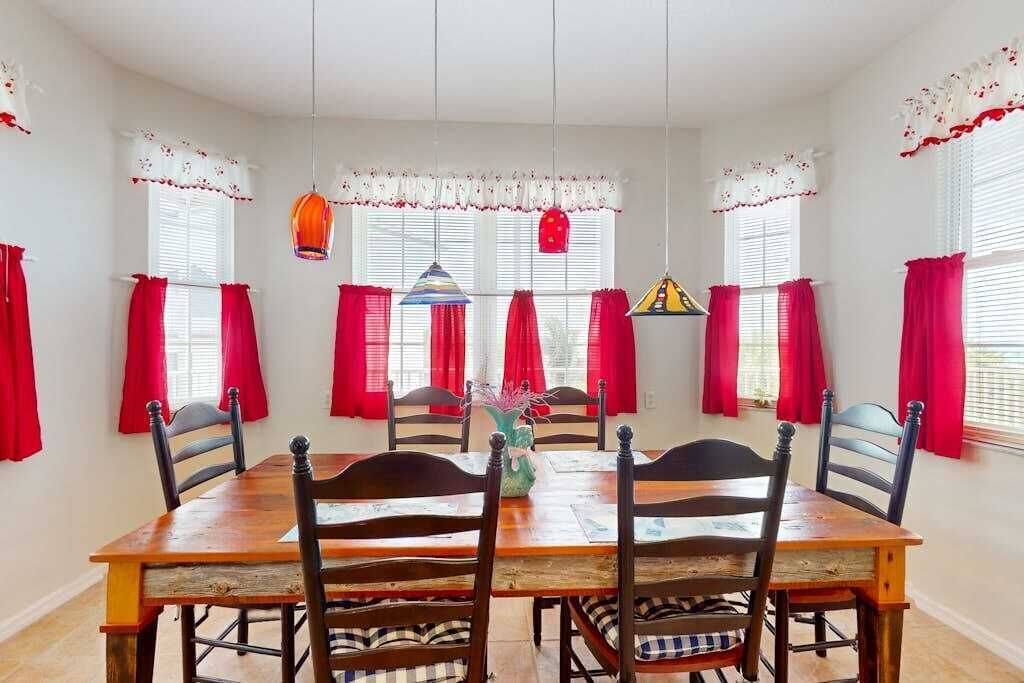 A dining room with a wooden table and chairs, red curtains, and hanging lamps.