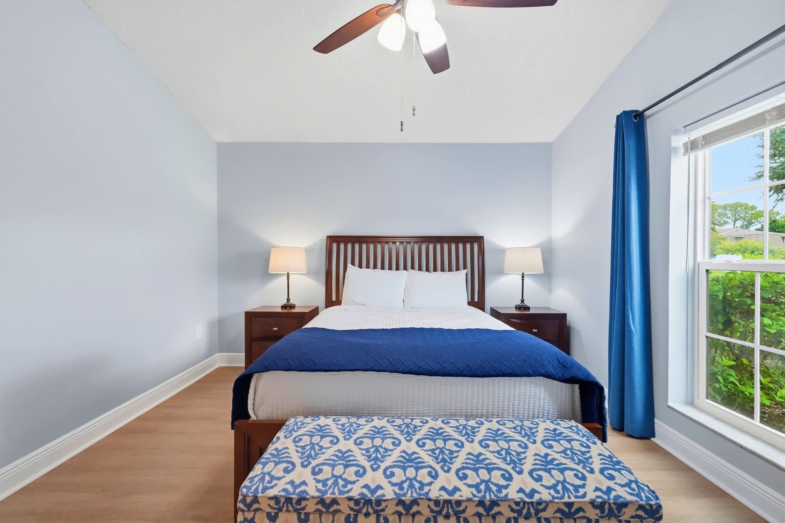 Une chambre à coucher avec un lit, deux tables de chevet avec des lampes, une fenêtre avec des rideaux et un ventilateur de plafond.