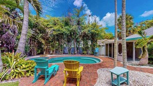 Zona de piscina amb terrassa de maó, dues cadiretes Adirondack, una taula petita i vegetació tropical al voltant.