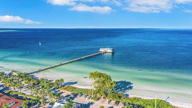 Vista costera con un muelle que se adentra en el océano, una zona de playa con coches aparcados y cielo despejado.