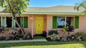 Una casa rosa con una puerta amarilla, contraventanas verdes y el número '4703' en la pared.