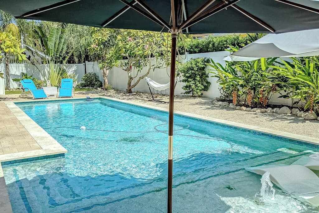A swimming pool with a tiled deck, two blue lounge chairs, and a white umbrella.