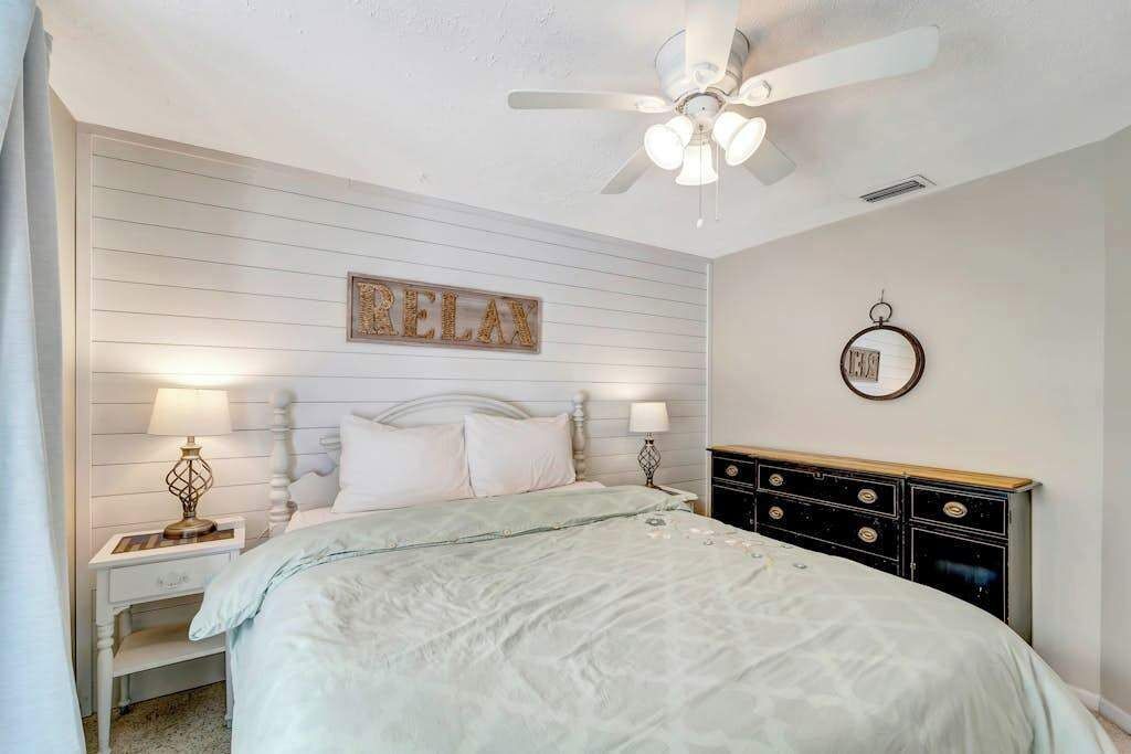 A bedroom with a bed, nightstands, a dresser, a ceiling fan, a wall-mounted mirror, and a 'RELAX' sign.