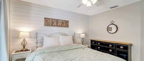 A bedroom with a bed, nightstands, a dresser, a ceiling fan, a wall-mounted mirror, and a 'RELAX' sign.