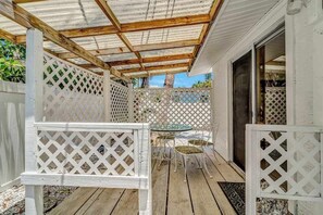 Ein überdachter Patio-Bereich mit Holzdeck, weißen Gitterelementen und einem Glastisch mit Stühlen.