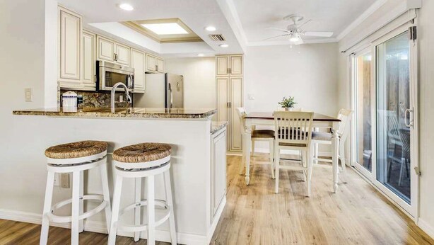 A kitchen with white cabinetry, a central island, two bar stools, a dining table with chairs, and a ceiling fan. - Charming Ground Floor Condo with Shared Pool, High-Speed WiFi, and Central AC (Holmes Beach)