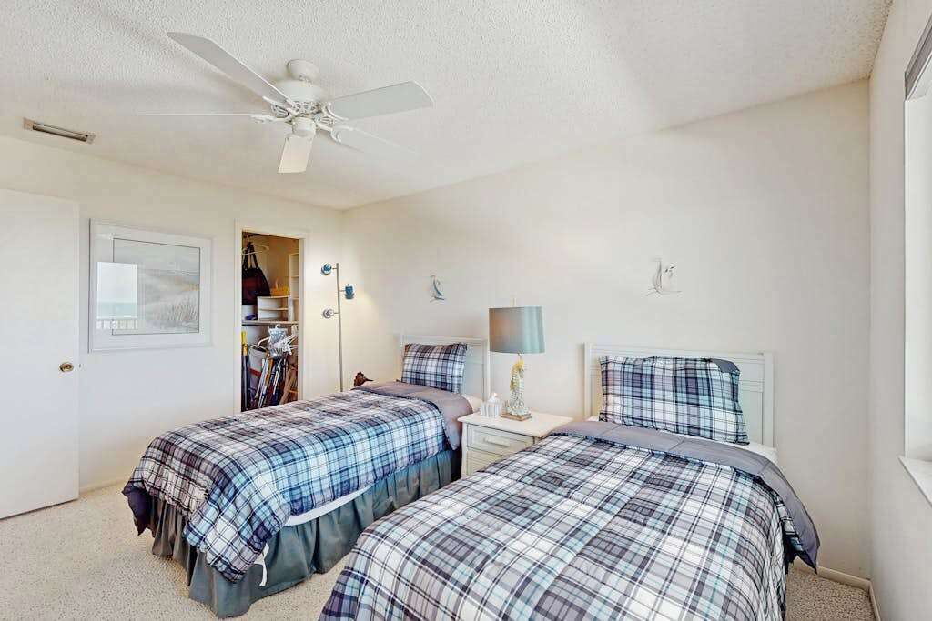Une chambre avec deux lits, un ventilateur de plafond, une table de chevet avec une lampe et une armoire avec des vêtements suspendus.