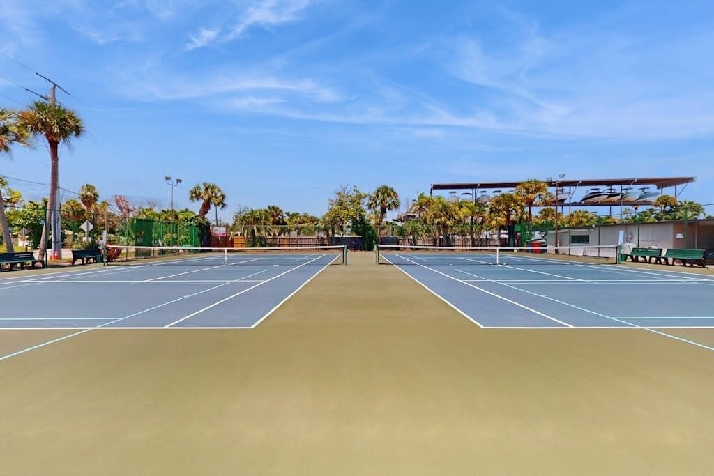 Een tennisbaan buiten met een net, omringd door palmbomen en banken.