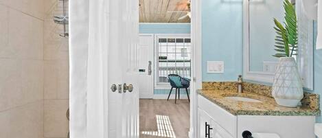 Baño con ducha, lavamanos y espejo. Hay una puerta que da a otra habitación.
