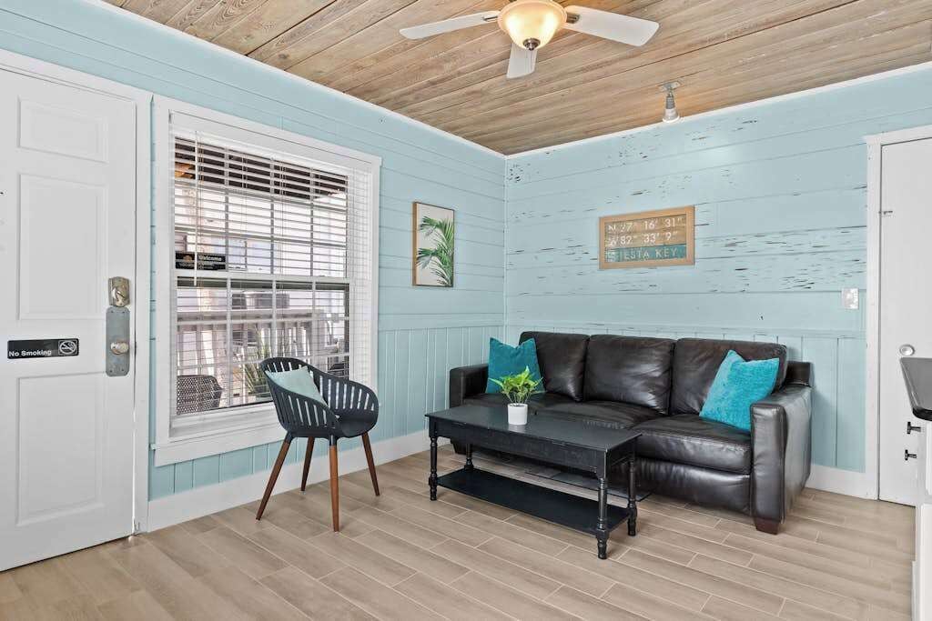 A room with a black leather sofa, a small table, a chair, a ceiling fan, and a window with blinds.