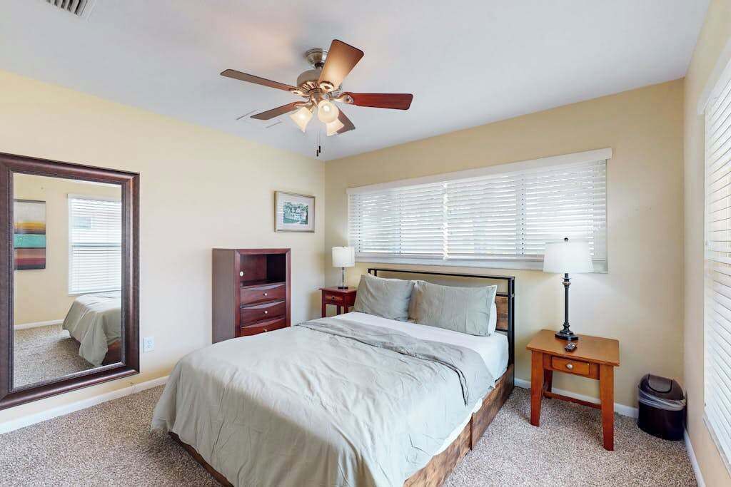 A bedroom with a bed, a ceiling fan, a window with blinds, a nightstand, a dresser, and a mirror.