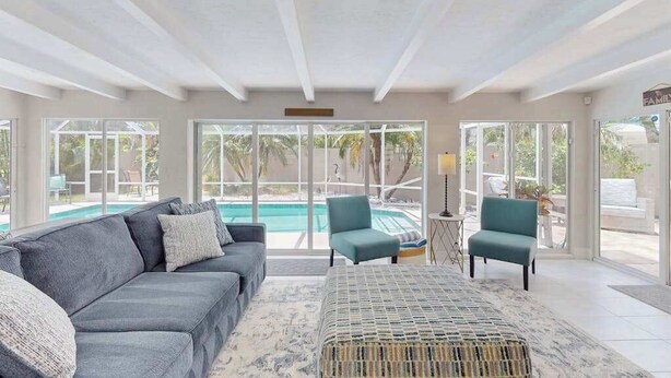 Ein großzügiger Wohnbereich mit grauem Sofa, Türkis-Armstühlen, einem gewebten Pouf und einem großen Fenster mit Blick auf einen Pool.