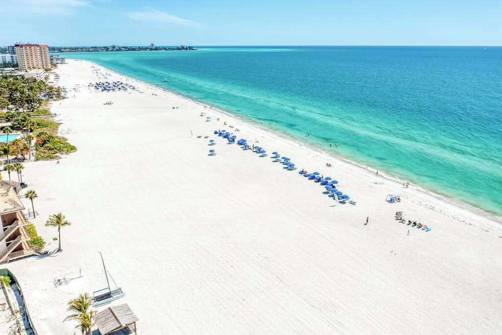 A beach with a row of blue umbrellas, a clear blue sea, and buildings in the background.