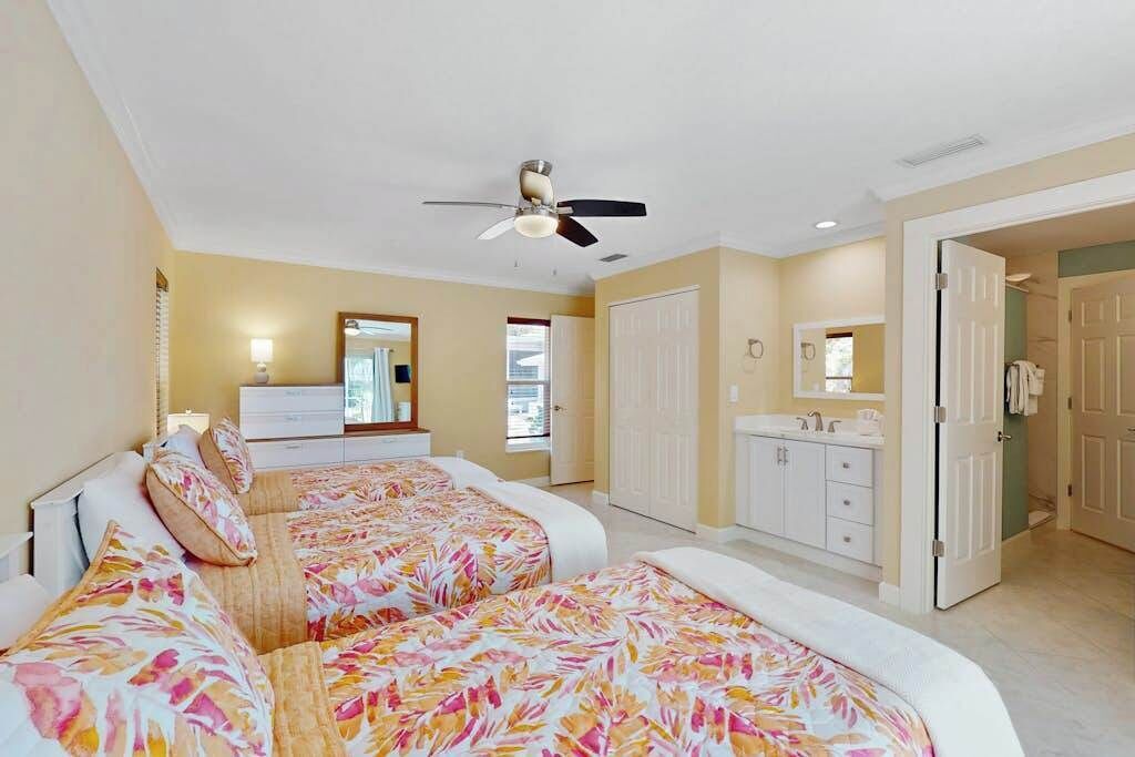 A bedroom with two beds, a ceiling fan, a bathroom with a sink and mirror, and a door leading to another room.