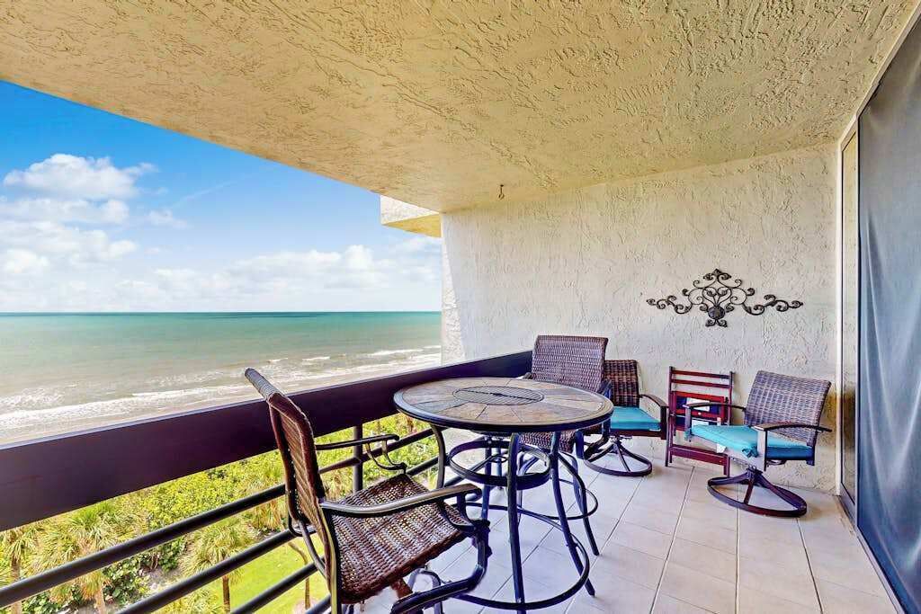 A balcony with a round table and four chairs, overlooking a beach.