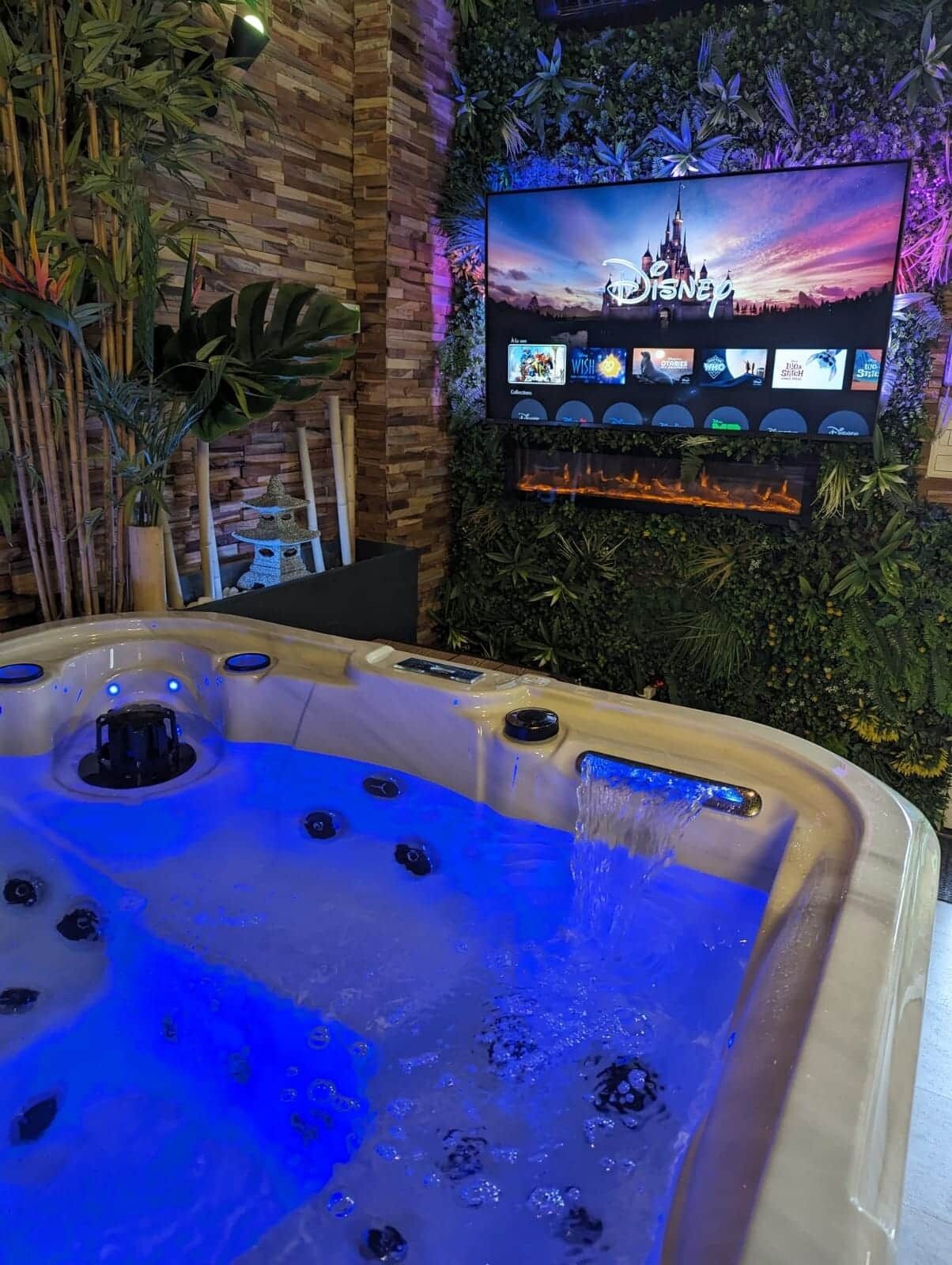 A hot tub with water jets, surrounded by indoor plants and a wall-mounted TV displaying Disney channels.