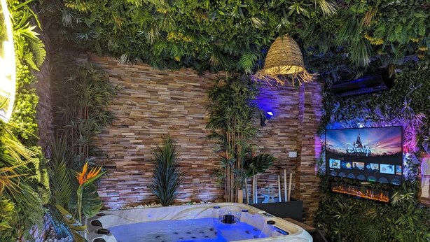 A hot tub surrounded by lush greenery and a stone wall, with a TV displaying Disney content.