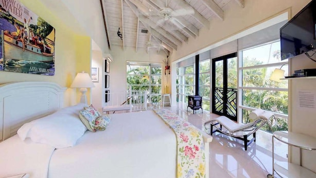 A spacious bedroom with a large bed, a sitting area with a bench, a television, and a view of the outdoors. - High-end home w heated private pool & spa, courtyard, & floor-to-ceiling windows (Anna Maria)