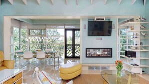 A spacious living room with a large TV, a fireplace, and a bookshelf with decorative items. - High-end home w heated private pool & spa, courtyard, & floor-to-ceiling windows (Anna Maria)