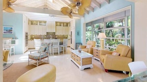 A spacious living room with a large window, a ceiling fan, and a kitchen in the background. - High-end home w heated private pool & spa, courtyard, & floor-to-ceiling windows (Anna Maria)