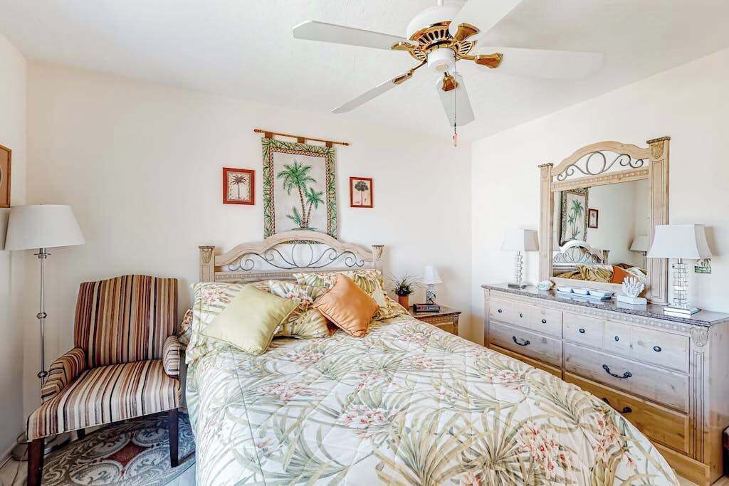 Una camera da letto con un letto, una sedia, una cassettiera, una ventola a soffitto e decorazioni alle pareti.
