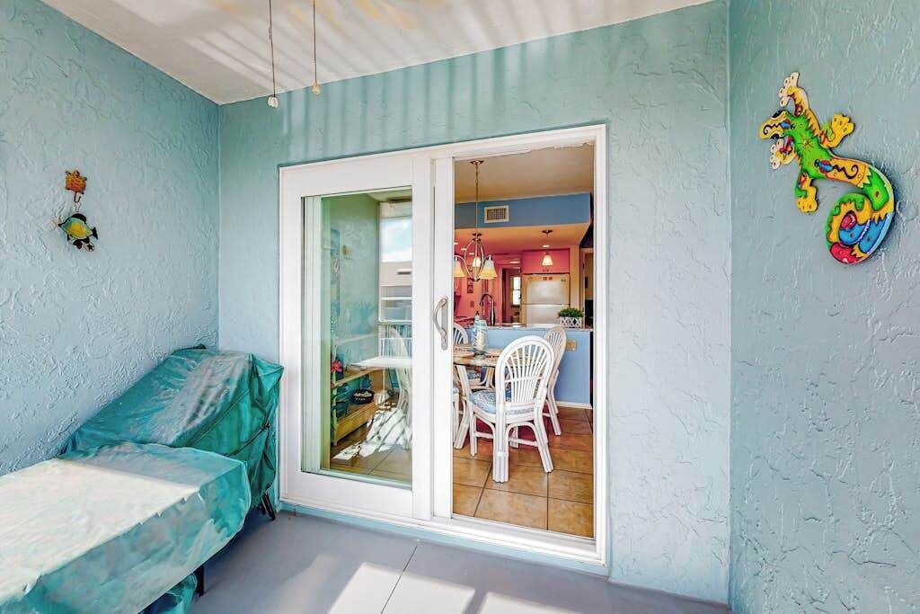 A room with a bed, a dining table with chairs, a kitchen area visible through a glass door, and a colorful wall decoration.