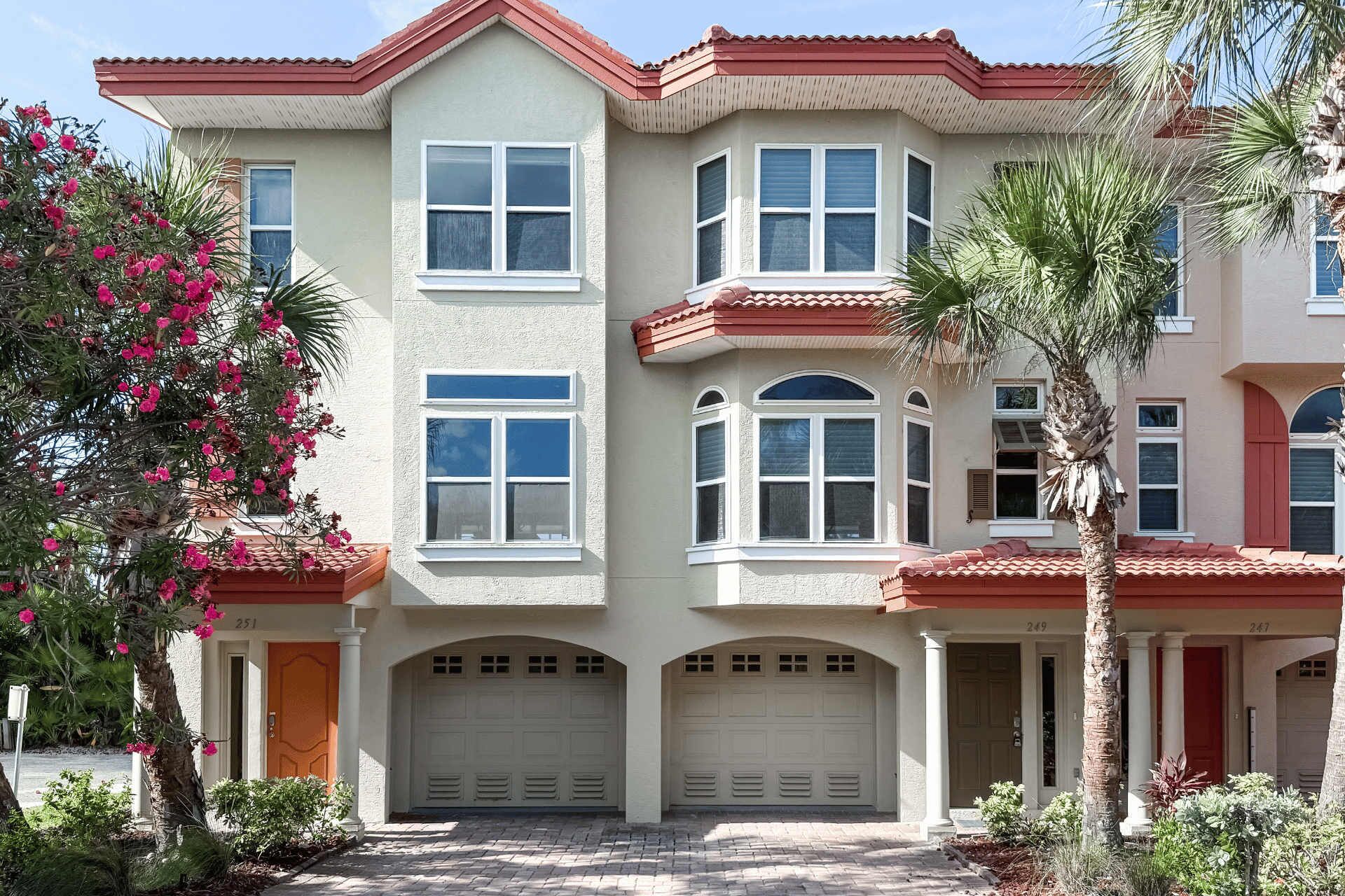 A multi-story residential building with a garage, a balcony, and a well-maintained garden with palm trees and flowering plants.