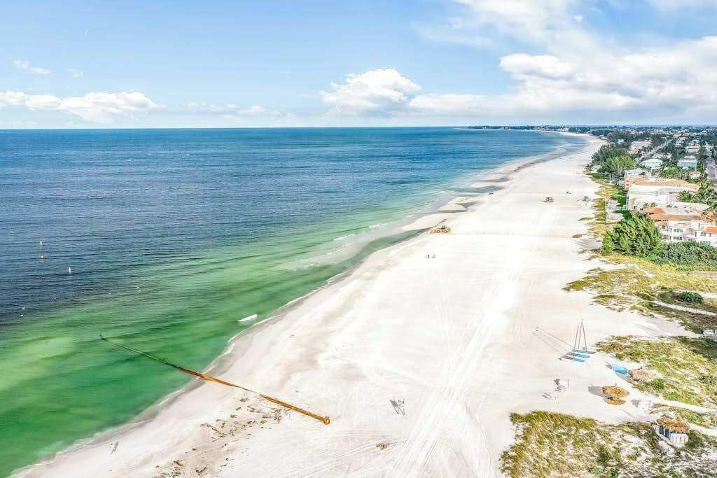 En strand med klart vatten, en sandstrand och byggnader längs kusten.