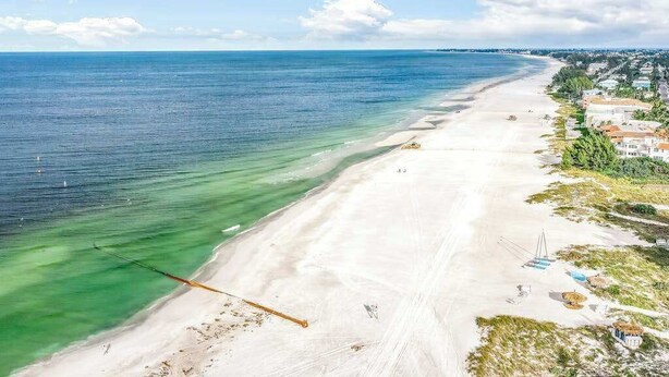 Ein Strand mit klarem Wasser, einem sandigen Ufer und Gebäuden entlang der Küste.