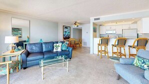 A living room with a blue sofa, a glass coffee table, and a dining area with wicker chairs. - Oceanfront condo with WiFi, & ocean views, walk to free trolley (Bradenton Beach)