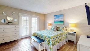 A bedroom with a bed, bedside tables, a bench, a dresser, a door, and a wall with mirrors and a painting. - Oceanfront condo with WiFi, & ocean views, walk to free trolley (Bradenton Beach)
